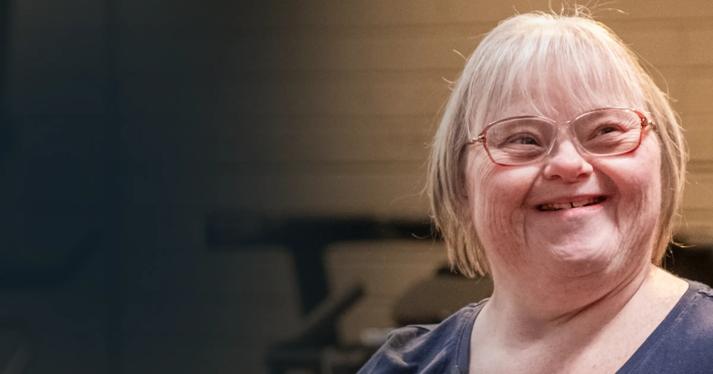 A woman with a joyful expression and glasses smiles warmly, positioned against a blurred indoor background, conveying happiness and positivity.